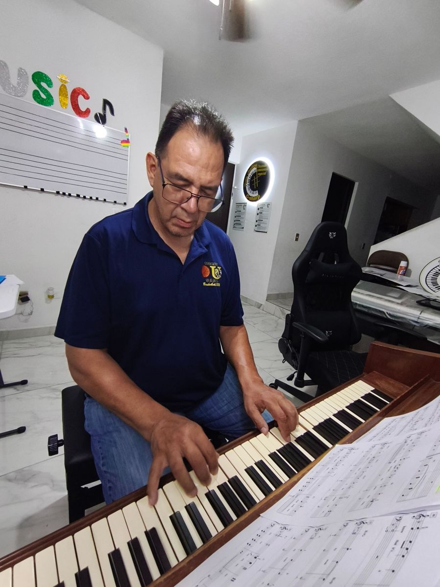 Un hombre adulto toca el piano con partituras al frente; al fondo se ven el pizarrón con pentagrama y el letrero del estudio