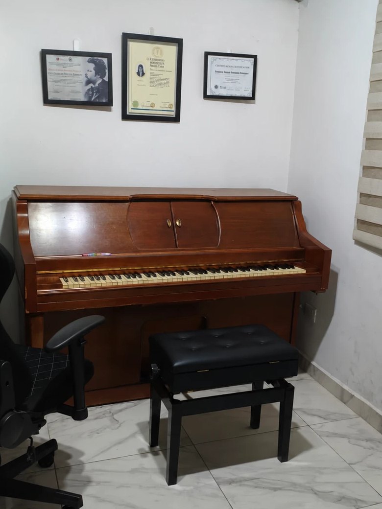 Piano vertical de madera con un banco negro acolchado frente al teclado; en la pared de arriba hay tres diplomas enmarcados