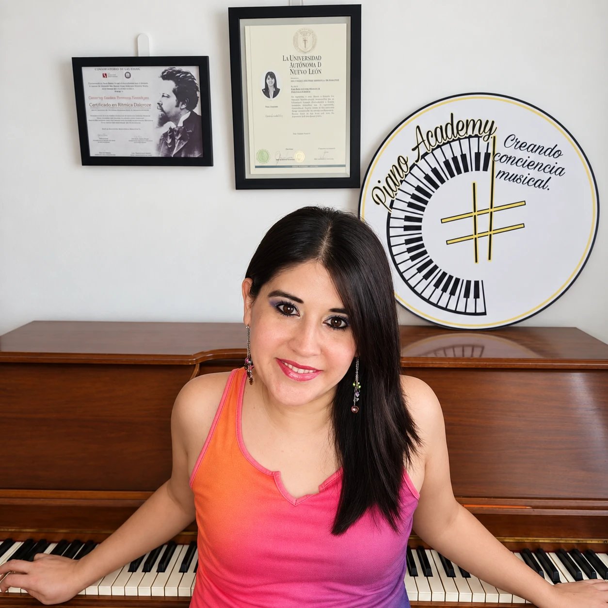 La profesora sonríe frente a un piano vertical; en la pared se ven dos diplomas enmarcados y el letrero del estudio