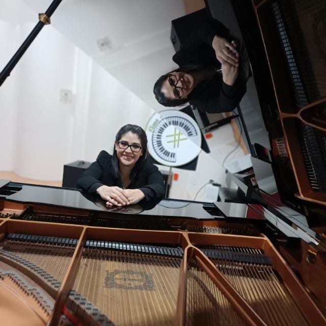 Deyanira Simental sonríe con las manos apoyadas sobre un piano de cola negro; al fondo se ve el letrero del estudio